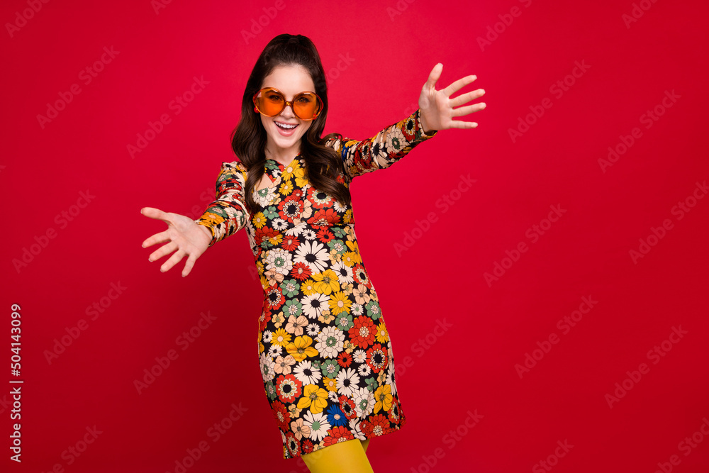 Photo of smiling woman posing on a red background, wearing 70s-style flower-print dress and round glasses, stretching her arms to camera.