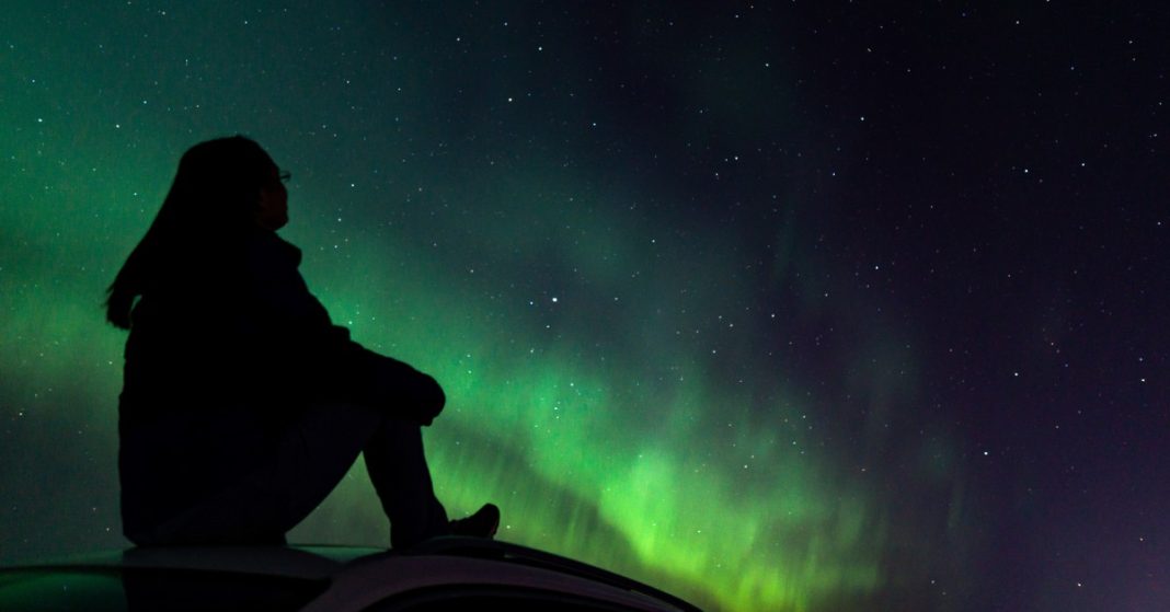 The silhouette of a woman sitting with her knees to her chest while looking up at the northern lights.
