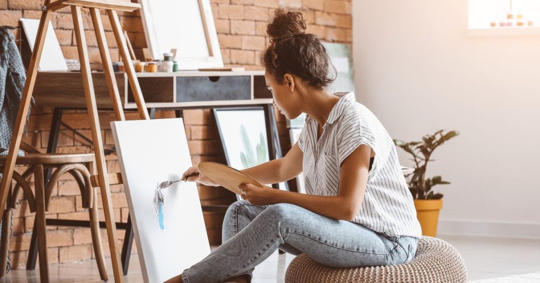 A young woman sitting on a cushion on the floor of her apartment as she focuses on painting on a canvas.