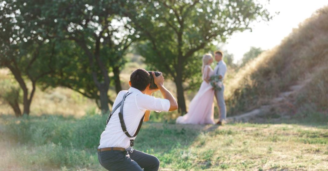 A wedding photographer taking a photograph of a bride and groom with luscious trees and fields in the background.